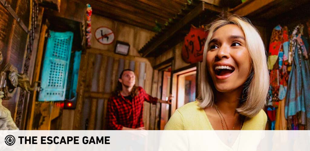 Image of two people in a warmly lit escape room, decorated with rustic clues and colorful wall hangings. A woman in the foreground laughs joyously while a man in the background looks on, smiling and engaged in the immersive puzzle-solving experience. Logo: The Escape Game.