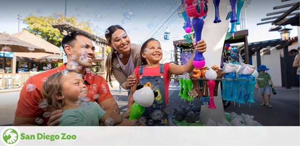 A joyful family at San Diego Zoo. A man, woman, and two children smile as they stand near a bubble machine, with glistening bubbles floating around them, capturing a moment of delight. The zoo's logo is visible at the bottom.