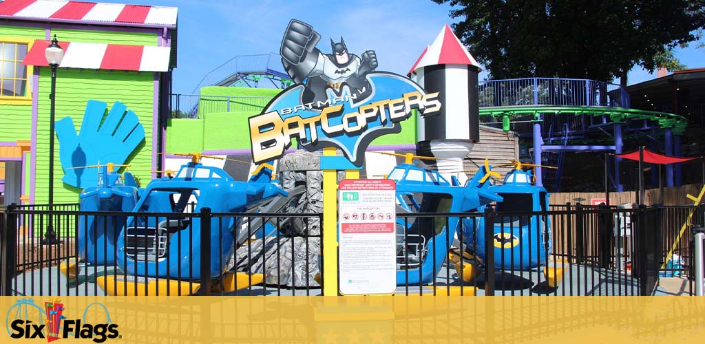 Image shows a colorful amusement park ride area under a bright blue sky. The centerpiece is a sign featuring the text 'BATCOPTERS' with a graphic of a superhero character above. The area includes blue helicopter-themed ride vehicles on a rail, surrounded by fencing, and nearby structures suggest a playful, vibrant atmosphere. The Six Flags logo appears in the corner.