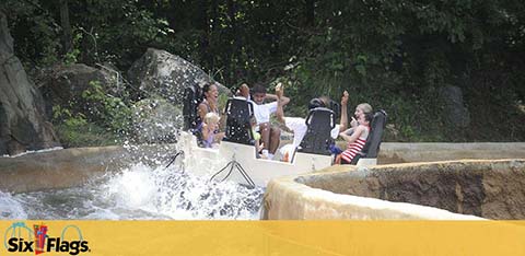 A group of excited people enjoys a thrilling water ride at Six Flags amusement park. They're splashed by water as their circular boat navigates a sharp turn surrounded by rocks and greenery. The Six Flags logo is visible at the bottom of the image. Fun and adventure await at the park for visitors.