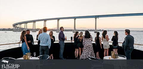 This image shows a group of people gathered on the deck of a boat enjoying an evening social event. The deck features modern styling with a sleek, dark-colored railing, and guests are dressed in casual to semi-formal attire, engaging in conversation and gazing out at the surrounding view. The atmosphere appears relaxed and convivial as the sky transitions to hues of pink and blue at dusk. In the background, a long, sweeping bridge spans the width of the photo, creating a striking silhouette against the fading light of the sky. The calm water reflects a gentle light, enhancing the serene maritime setting. Prominent on the image is the bold logo for "FLAGSHIP."

At GreatWorkPerks.com, we ensure that your experiences are not only memorable but also affordable. Take advantage of our latest offers to get the greatest savings and lowest prices on tickets for your next marine adventure.