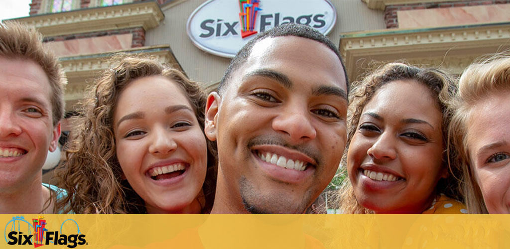 A diverse group of five joyful friends takes a close-up selfie in front of a 'Six Flags' theme park entrance, displaying wide smiles, evidently having a great time. The Six Flags logo is prominent in the background.