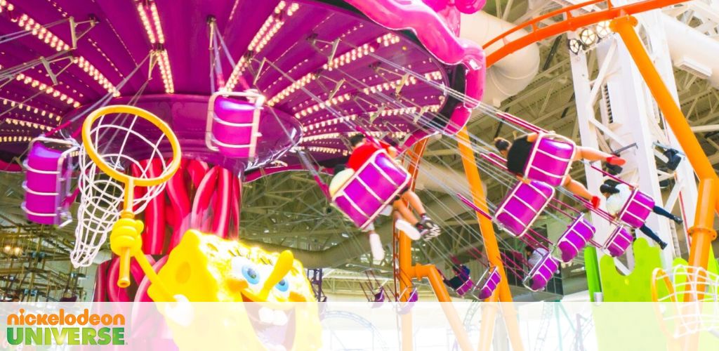 Image features a vibrant amusement park attraction with purple basket seats suspended from a rotating center. Below, a giant character with a net indicates a playful theme. Overhead, the structure's bright orange framework adds contrast to the indoor setting.