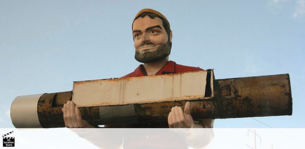 A large statue of a smiling man with a beard hoists a long, horizontal object over his shoulder against a blue sky. He wears a red shirt and cap and exhibits a caricatured, friendly appearance. The object seems like a vintage advertisement or sign.