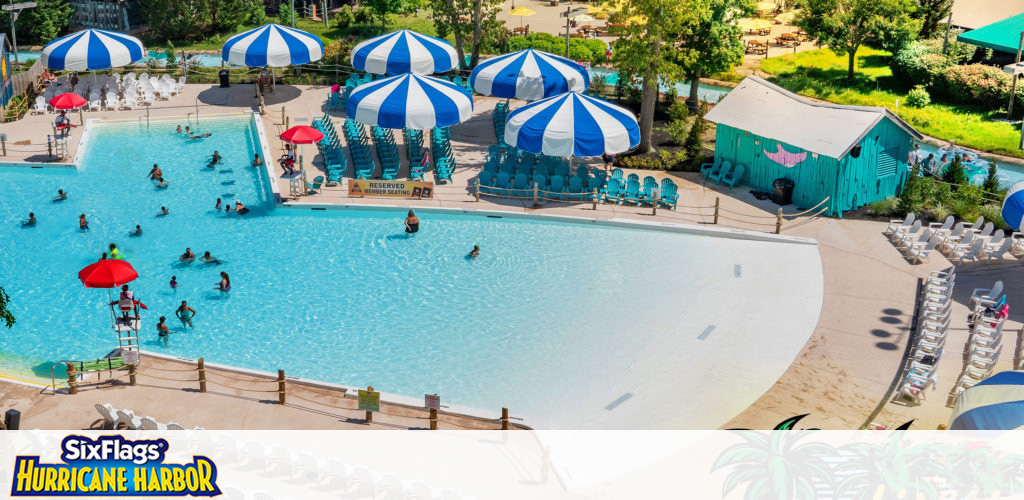 Above view of a vibrant water park at Six Flags Hurricane Harbor. Blue and white striped umbrellas dot the area, shading lounge chairs. Guests enjoy the spacious wave pool under a sunny sky. A green shack and palm trees add to the tropical theme.