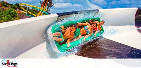 Four people are enjoying a water slide ride at Six Flags, with expressions of excitement. They're in a blue inflatable raft on a curved white slide, with a clear blue sky and another slide in the background. The Six Flags logo is visible.