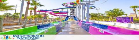 Image of Schlitterbahn Waterpark showing colorful water slides against a blue sky. Trees and structures surround the slides, with no people visible, emphasizing the vibrant slides and sunny weather.