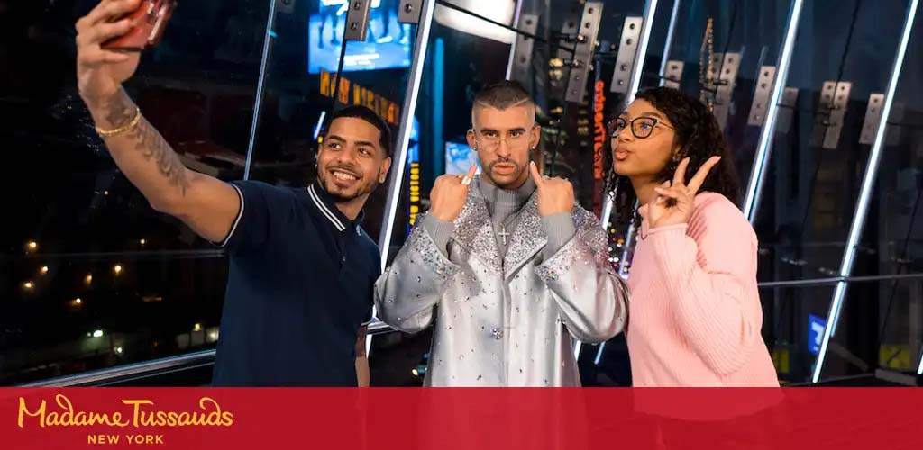 Three people are posing for a selfie in a lively setting shown by the label Madame Tussauds New York. A man on the left extends his arm to capture the moment, a wax figure in the center is styled in a sparkling outfit mimicking a thumbs-down gesture, and a woman on the right flashes a peace sign with a smile.