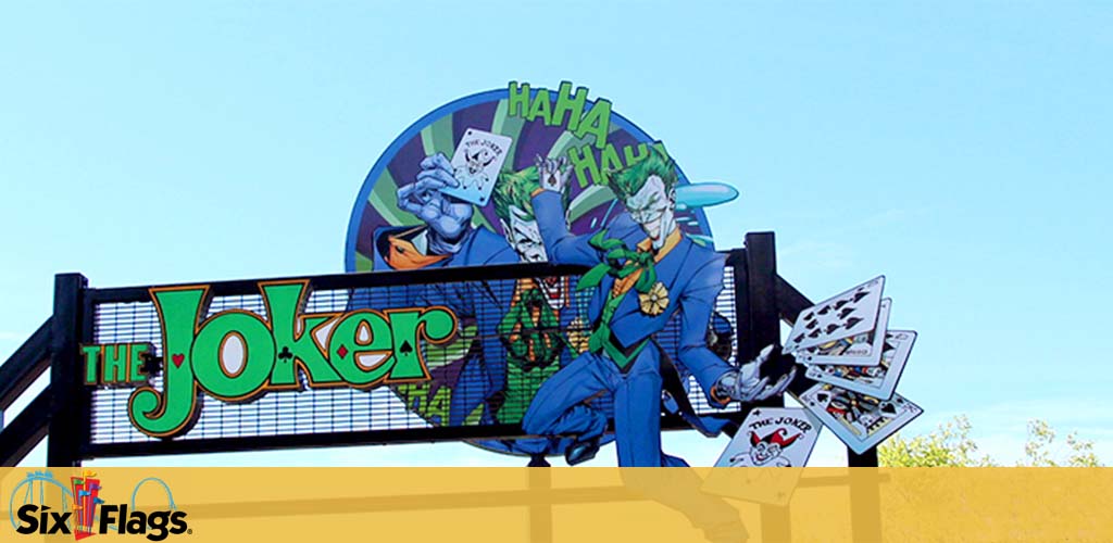 Entrance sign for 'The Joker' ride at a Six Flags theme park. It features a large oval with The Joker, a comic book character, in a mischievous pose, holding a fan of cards, with 'HAHAHA' in the background. The sign has large, stylized text spelling 'The Joker' beneath the graphic. The Six Flags logo is visible in the lower left corner against a clear blue sky.