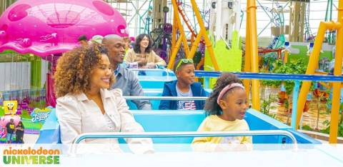 Image of a joyful family on a theme park ride at Nickelodeon Universe. Two adults and two children are smiling and looking excited as they sit in a blue roller coaster car. A large pink character balloon and colorful amusement park structures are in the background.