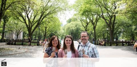 Three people are smiling and holding ice cream cones in a tree-lined park on a sunny day. The area is bustling with activity and visitors enjoy the greenery and open space.