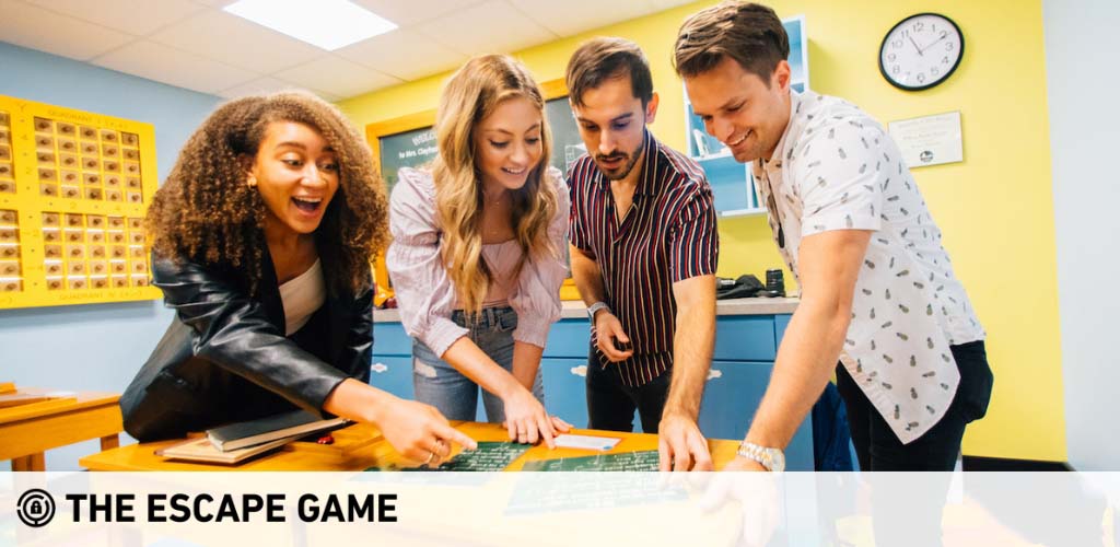 Four people stand around a table in an escape room, collaborating on a puzzle. A woman with curly hair shows excitement, while the others are focused on finding clues. The room is brightly lit with a lively atmosphere. The Escape Game logo is featured in the lower left corner.