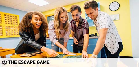 Four people are gathered around a table in a brightly colored room, intently working together on a puzzle or game. They appear focused and engaged in teamwork. The setting suggests an escape room activity. Below the image is the logo for The Escape Game.