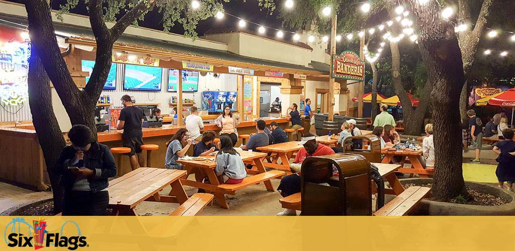 Outdoor dining area at night with wooden picnic tables where people are seated eating and socializing. There's a walk-up food counter with a menu display and staff serving. Trees with lights add ambiance, and a Six Flags logo is visible in the upper left corner.