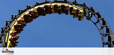 Riders are inverted on a roller coaster loop against a clear blue sky. Below the loop, the Six Flags logo is visible, hinting at the thrilling amusement park experience. The guests appear secured in their seats, adding an element of safety to the excitement.