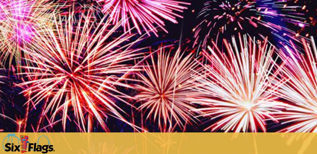 Vibrant fireworks fill the sky with bursts of pink, red, and white, set against a night backdrop above a Six Flags logo at the bottom. The display conveys a celebration or event at the amusement park.