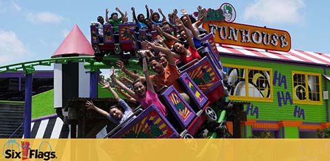 Visitors enjoy a thrilling ride at a Six Flags amusement park on a sunny day. The brightly colored roller coaster is in a steep drop, with riders' arms up in excitement. In the background is a vibrant funhouse with playful designs and the Six Flags logo prominently displayed.