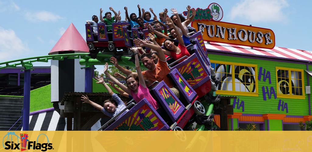 Thrilled riders raise their arms on a colorful roller coaster at Six Flags, with the Joker's Funhouse in the background under a clear blue sky.