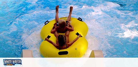 Two individuals are on a yellow water raft sliding down a blue, foamy water slide. They appear to be holding onto handles, and one raises their arms in excitement. The slide seems to be at an outdoor aquatic park. There's a logo that reads 'White Water' at the bottom.