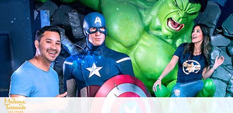 A man and a woman are posing with life-size figures of Captain America and the Hulk at Madame Tussauds. The man, to the left, smiles beside Captain America, who is in full costume holding a shield. The woman, to the right, appears excited, imitating the Hulk's roar. Background resembles a demolished wall, enhancing the superhero theme.