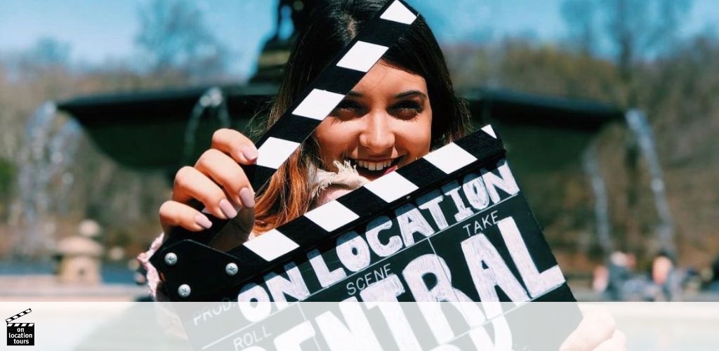 A smiling woman holds a clapperboard with the words  ON LOCATION TOURS  written on it. She appears to be outdoor at a sunny location with trees and a fountain in the background, suggesting an upbeat, entertaining setting possibly connected to filming or touring.