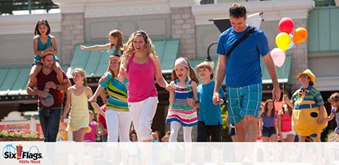 Group of happy visitors at a theme park, walking and enjoying a sunny day. There's a mix of adults and children; some are holding hands. One kid with balloons adds a festive touch to the scene. A Six Flags logo is visible, indicating the location.