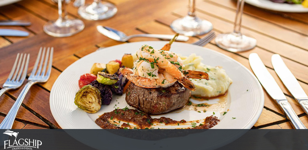 Image description: A gourmet meal is elegantly presented on a white circular plate, which rests on a wooden tabletop. At the center of the plate is a well-seared steak, topped with two grilled shrimps and garnished with green herbs. To the side, there is a dollop of creamy mashed potatoes and an assortment of roasted vegetables including Brussels sprouts, cherry tomatoes, and possibly some pieces of roasted beetroot, providing a vibrant array of colors. The table is set with two forks on the left, and on the right, two knives and a wineglass, suggesting a fine dining experience. Accompany fun-filled memories with the taste of luxury cuisine, all while enjoying the savings as GreatWorkPerks.com consistently offers the lowest prices on tickets and dining experiences.