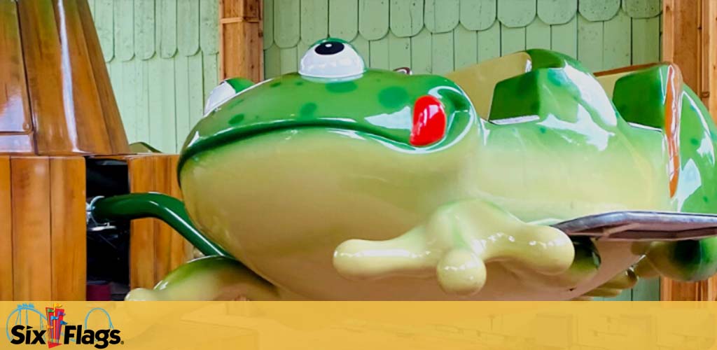 Image shows a close-up of a large, vibrant green frog sculpture with white and red detailing, likely part of a theme park attraction. The frog has a friendly appearance with big bulging eyes looking to the side, perched as if ready to hop. In the background, there is what appears to be a wooden fence, indicating an outdoor setting. The logo of Six Flags is visible in the corner, suggesting the location.