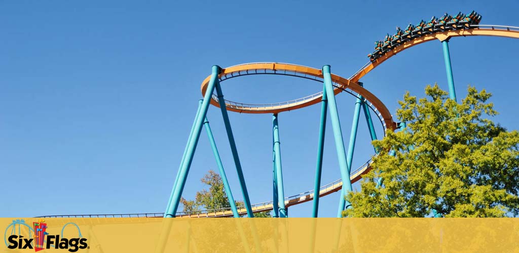 Image shows a roller coaster with orange tracks and teal supports against a clear blue sky. A filled coaster train crests a curved track. Trees with green foliage are visible below the track. The Six Flags logo is in the bottom left corner on a gradient yellow bar.