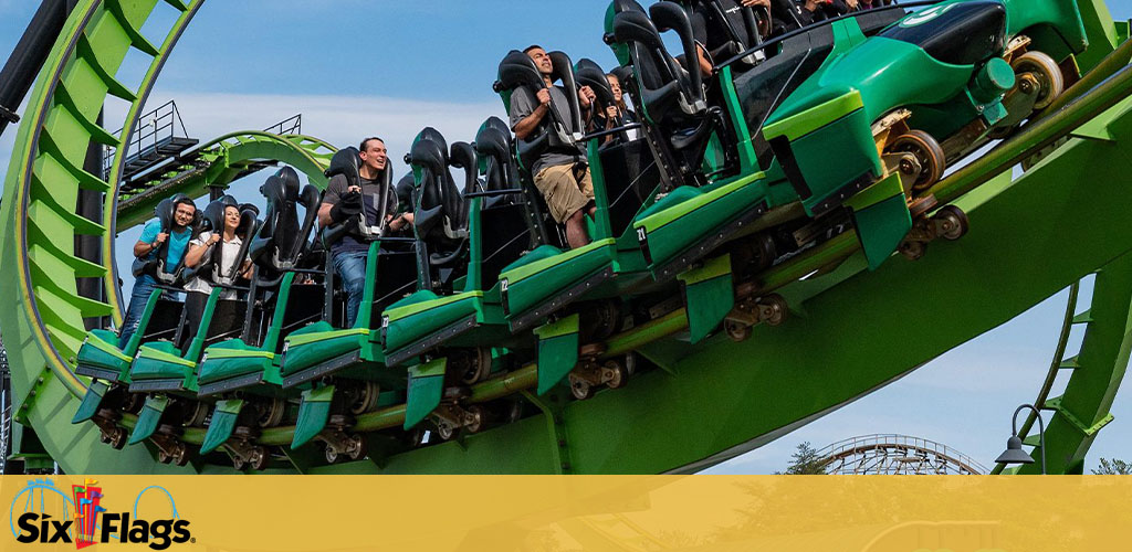 Image shows a roller coaster in mid-ride with excited passengers. The coaster is green with a Six Flags logo displayed, indicating the theme park’s thrilling atmosphere. Passengers appear to be enjoying the exhilarating experience.