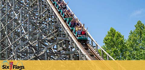 Visitors enjoy a thrilling ride on a wooden roller coaster at Six Flags, ascending a steep track with excitement under a clear blue sky. Lush trees line the background, and the Six Flags logo appears prominently.