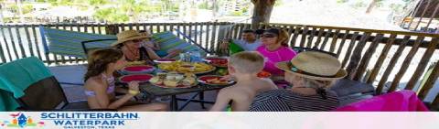 People enjoy a meal in a cabana at Schlitterbahn Waterpark. Guests are sitting around a table with plates of food, engaging in conversation. The cabana has a thatched roof, and the atmosphere seems relaxed with several individuals wearing sun hats.