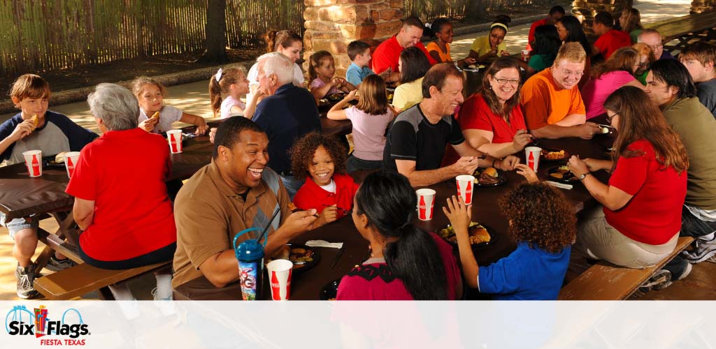 Group of people of various ages enjoying meals at picnic tables. Everyone is casually dressed, with smiles and engaged in conversation. The setting appears to be a shaded outdoor area, with the logo of Six Flags Fiesta Texas visible, suggesting a theme park environment.