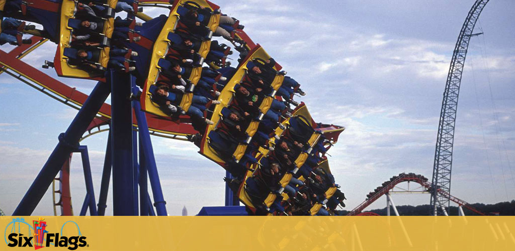 Photo of a roller coaster ride in action at a Six Flags amusement park. Passengers in secure seats are upside down mid-loop, expressing excitement. The coaster is multicolored with a clear sky background. The Six Flags logo is visible on the lower left.