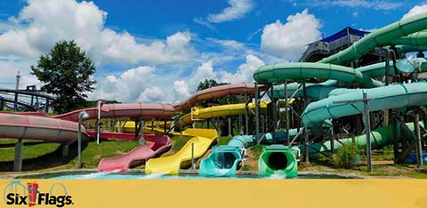 Image of a colorful array of water slides at a Six Flags theme park under a blue sky with puffy clouds. The foreground shows the end of slides with splashes and the entrance to green, yellow, and pink tube slides, signaling fun and excitement.