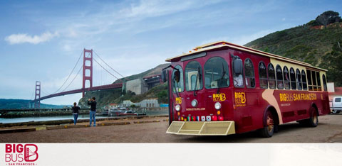 Description: The image features a vibrant red and gold "Big Bus San Francisco" tour bus parked on a paved surface near a coastal area. The iconic Golden Gate Bridge is visible in the background, spanning the horizon with its towering red suspension structures dipping into the rolling green hills on either side. To the left of the bus, two individuals are seen, with one seemingly capturing a photograph of the other standing with the bridge in the background. The sky is partially overcast, yet daylight is ample, illuminating the scene with natural light.

At GreatWorkPerks.com, we are committed to helping you explore the wonders of San Francisco with the added benefit of unbeatable savings – find the lowest prices on tickets for your next adventure with us!