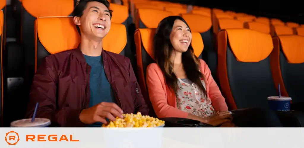 A man and a woman are enjoying a movie while seated in a cinema with bright orange seats. The man, to the left, is laughing and holding a popcorn container, while the woman, to the right, smiles widely. A beverage is visible by her side. The Regal Cinemas logo is displayed in the lower right corner.