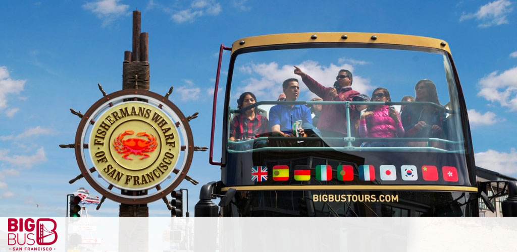 This image depicts a group of joyful tourists on the upper deck of a Big Bus Tours double-decker bus, with a clear blue sky overhead. On the left, the iconic circular Fisherman's Wharf sign of San Francisco, featuring a crab design in the center and encircled with yellow lettering, is prominently displayed. The bus is adorned with flags representing various countries, signifying the international appeal of the city's attractions. The tourists appear engaged and are pointing at sights, capturing photographs, and enjoying the open-air experience. At GreatWorkPerks.com, we're committed to providing you with the lowest prices and great savings on tickets to similar memorable experiences.