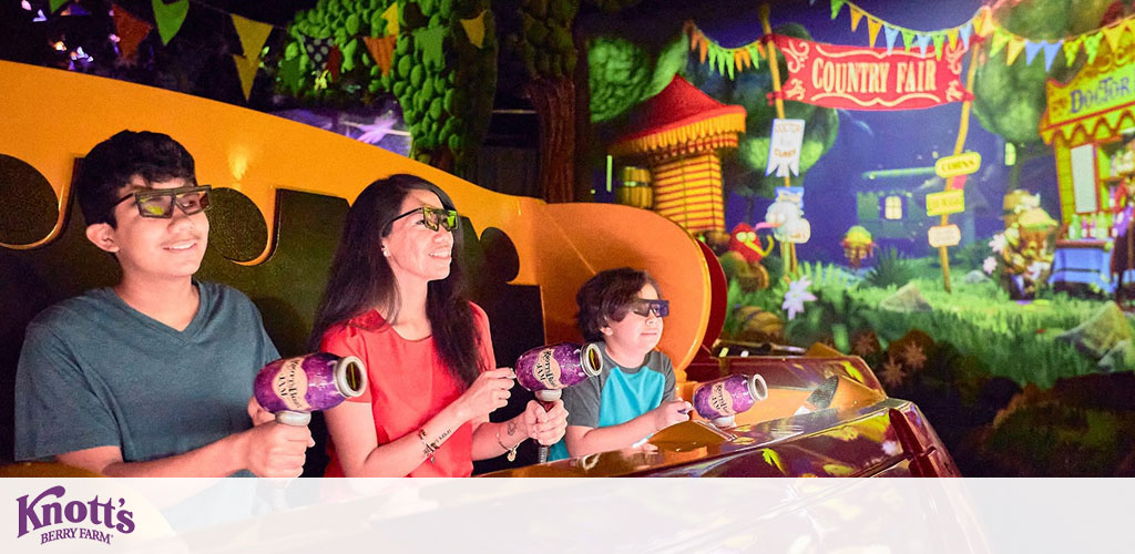 Three people are seated in a bright yellow ride vehicle, each holding a blaster, wearing 3D glasses, and aiming at targets in a vibrant, fairy tale-themed shooting gallery with signs reading 'Country Fair'. A Knott's Berry Farm logo is visible in the corner.