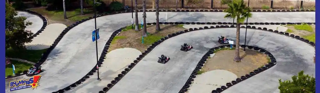 This image showcases an outdoor go-kart racing track during daytime. The track twists and turns, forming a serpentine pattern, bordered by a series of black tires with white tops that line the edges and separate the lanes. A single palm tree stands as a focal point in one of the track's internal divides, surrounded by an area of sand and grass. Three go-karts, each with a single occupant, are seen engaging in the activity. The leading kart is in motion on the left side of the track while two other karts follow behind on the curving path to the right. The surrounding area is well-maintained with visible landscaping and a clearly marked pedestrian path to the side.

Enhance your experience with us and get ready for an exhilarating ride at GreatWorkPerks.com, where our commitment to savings means offering you the lowest prices on tickets for memorable adventures!