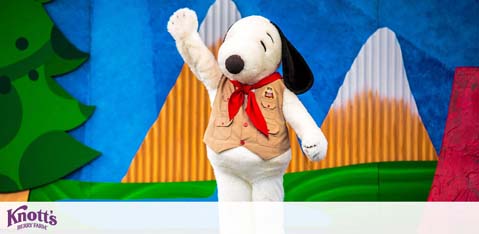 Image shows a costumed character resembling Snoopy from the Peanuts comics. It's waving happily, dressed in a beige scout's uniform with a red neckerchief. Snoopy is on stage with a backdrop featuring cartoon-style mountains. The Knott's Berry Farm logo is visible, indicating the location or event. The setting appears cheerful and kid-friendly.