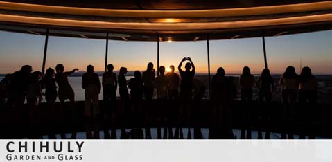 This image features a silhouette of people gathered inside a room with panoramic windows, against the backdrop of a sunset. The sun is centered and partially obscured by a figure, creating a focal point. The room's ceiling curves overhead, and the bottom of the image has text that reads 'CHIHULY Garden and Glass'.