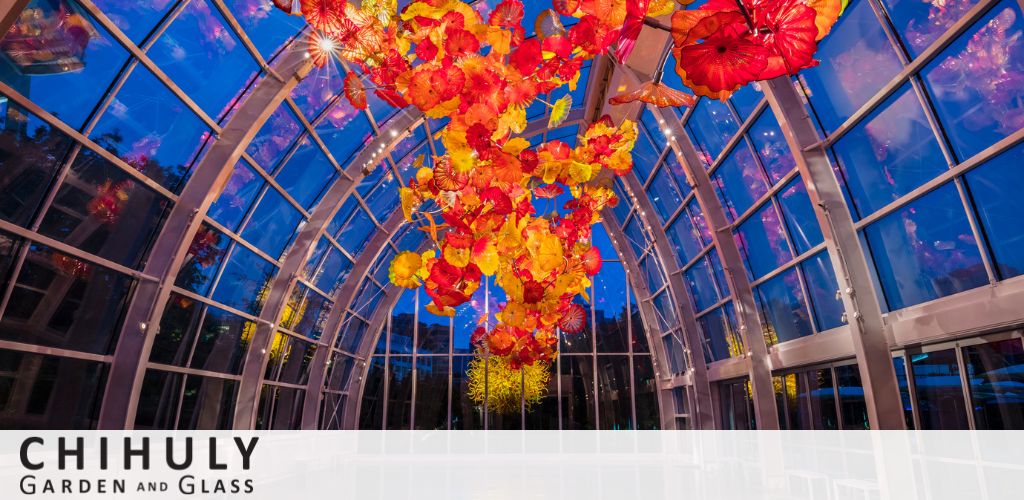 Vibrant glass sculptures by Chihuly hang from the ceiling in a glasshouse. The artwork features an array of fiery red, orange, and yellow colors reminiscent of flowers and sea creatures, contrasting with the blue sky visible through the transparent roof. The Chihuly Garden and Glass logo is on the bottom left.