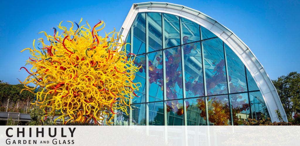 This image shows a large, vibrant yellow and red glass sculpture resembling a burst of sea anemones in front of a modern, curved glasshouse under a clear blue sky. Text on the image reads 'Chihuly Garden and Glass,' indicating the setting. The sculpture offers a striking contrast to the neatly manicured greenery partially visible around it.