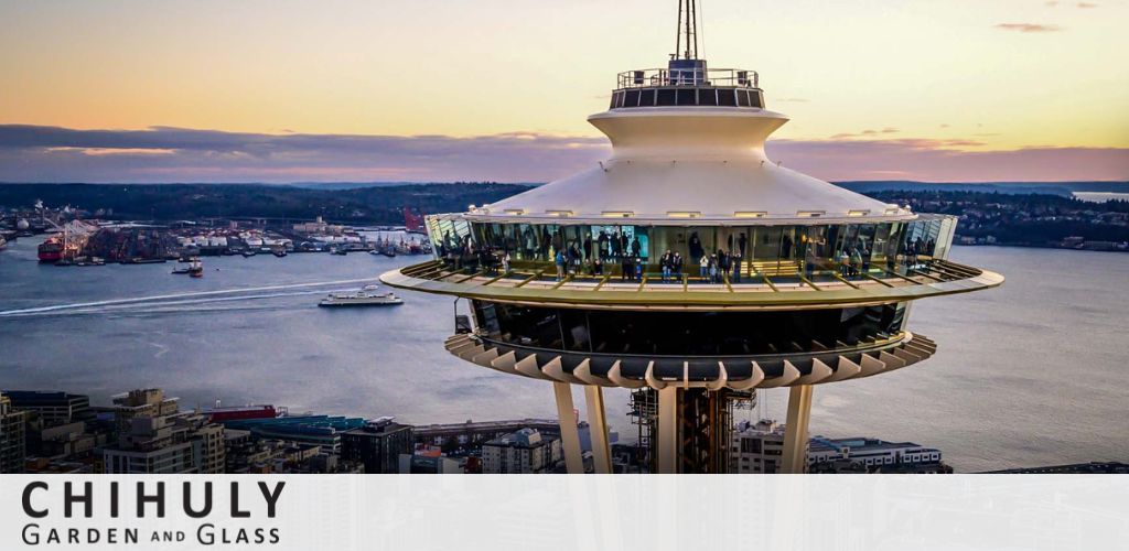 Image shows a scenic view of a famous tower with its observation deck highlighted against a dusky sky, overlooking a cityscape and waterway with boats. The tower's lights are on, offering a warm glow. Text indicates an association with Chihuly Garden and Glass.