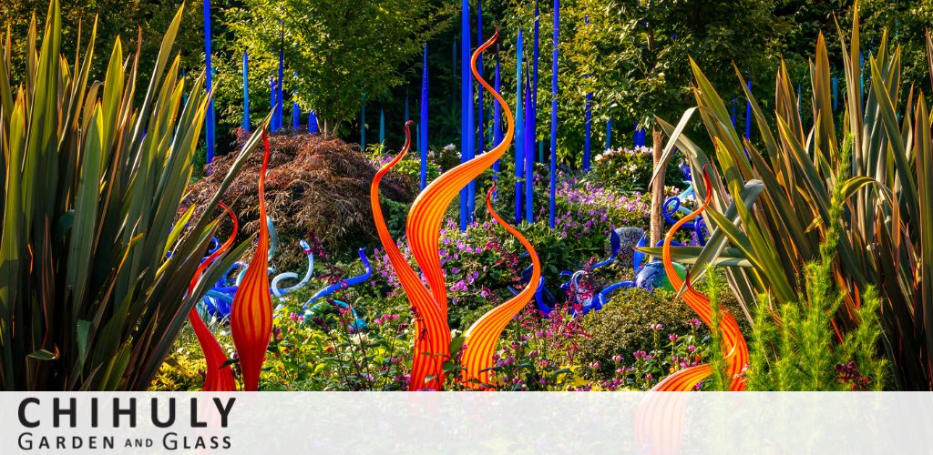 Image of Chihuly Garden and Glass displays with vibrant glass sculptures intertwined among lush greenery. Tall blue and orange spiraled forms rise above a variety of plants under sunlight.