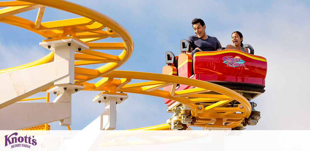 Image shows two people enjoying a ride on a yellow and orange roller coaster named Coast Rider at Knott's Berry Farm. They seem thrilled, with the sky as the backdrop. The front cart is prominently red with the ride's name on it. Knott's logo is visible below.