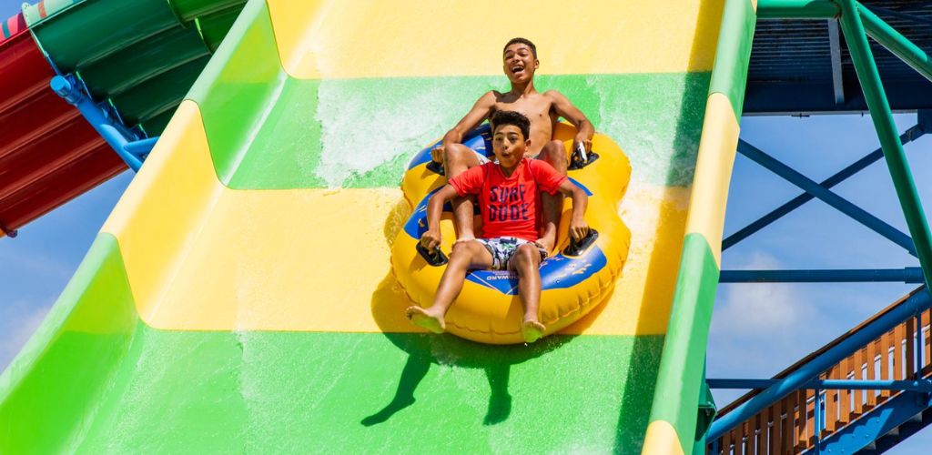 Two people joyfully slide down a colorful water slide on a raft.