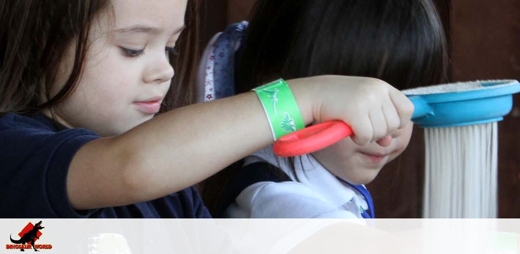 Two young children are focused on a science educational activity. The child in the foreground is using a red tool to handle a blue object, possibly part of an experiment or craft. They wear a green wristband with designs. A logo with a dinosaur and the text 'Dinosaur World' is in the lower left corner. The setting suggests a playful and learning environment.