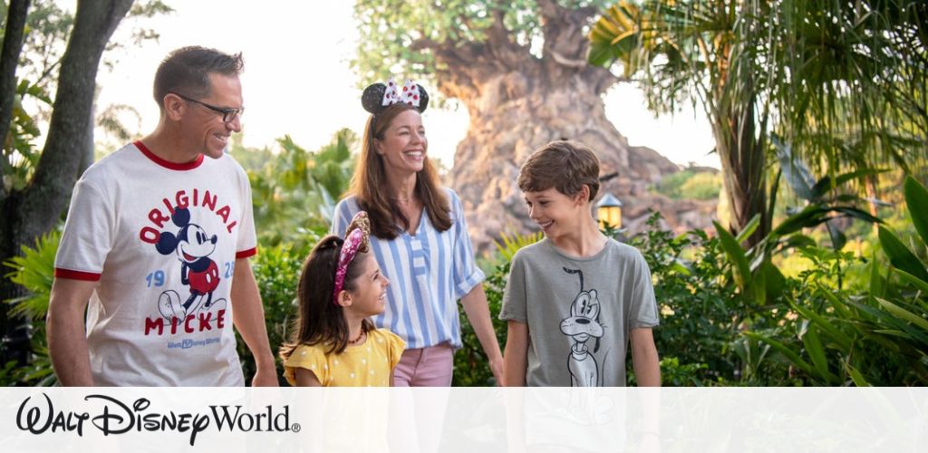 A family of four enjoys a day at a theme park, standing amidst lush greenery. The father wears an Original Mickey shirt, the mother wears Minnie Mouse ears and a striped shirt, the son wears a Pluto shirt, and the daughter in a yellow polka dot dress wears a sparkly headband. They all smile and look at each other, with a large tree in the background.